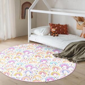 May include: A round, watercolor-style rug featuring a repeating pattern of colorful rainbows and polka dots on a white background. The rug has a pink border and is placed on a light wood floor. The rug is designed for a child's room.