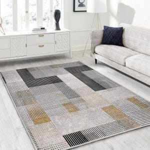 May include: A gray and white geometric patterned area rug with black and gold accents. The rug has a modern design with a mix of stripes and squares.