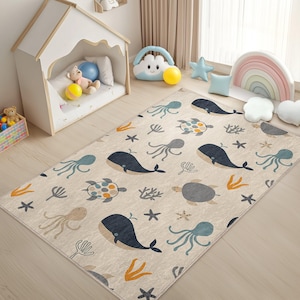 May include: A beige rectangular rug with a sea life pattern. The rug features illustrations of whales, octopuses, turtles, starfish, and coral in shades of blue, gray, and orange. The rug is placed on a light-colored wooden floor.