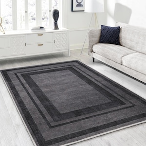 May include: A gray area rug with a multi-layered border design. The rug has a dark gray center and a lighter gray border with a subtle sheen.