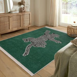 Puede incluir: Una alfombra rectangular verde con borde blanco. La alfombra presenta un gran diseño de leopardo estilizado en tonos grises y negros. El leopardo está tumbado, con el cuerpo estirado sobre la alfombra.