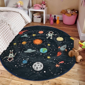 Könnte beinhalten: Ein runder, dunkelblauer und schwarzer Spielteppich mit Weltraum-Motiv und blauem Rand. Der Teppich zeigt Illustrationen von Planeten, Sternen, Raketen und Astronauten.