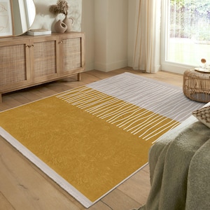 May include: A mustard yellow and grey area rug with a white abstract pattern. The rug has a white border and is laid out on a wooden floor.