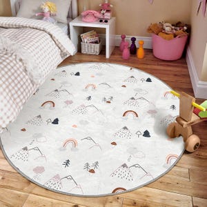 Op de afbeelding: Een ronde, witte speelmat met een grijze rand heeft een herhalend patroon van bergen, regenbogen en bomen. De mat is perfect voor de speelkamer of slaapkamer van een kind.