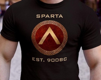 Puede incluir: Camiseta negra con la palabra "SPARTA" sobre un emblema circular. El emblema presenta un centro rojo con un galón dorado y un borde dorado de clave griega. El texto "EST. 900BC" está debajo.
