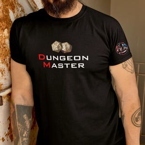 Op de afbeelding: Een zwart T-shirt met een witte afbeelding van twee dobbelstenen en de tekst "DUNGEON MASTER" in rode en witte letters. Het shirt heeft een klein patch op de mouw met een afbeelding van een krijger.