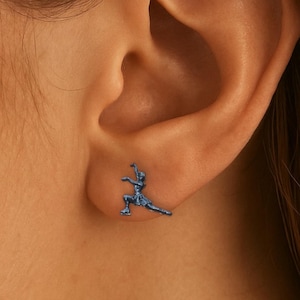 May include: A blue stud earring depicting a person in a yoga pose is shown on an ear. The earring is a detailed miniature sculpture, highlighting the figure's form and posture. The earring is a small, elegant accessory.