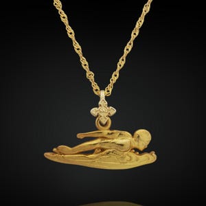 Può includere: Una collana con pendente dorato raffigurante una figura stilizzata sdraiata su una base curva. Il pendente è sospeso a una delicata catena d'oro. Lo sfondo è un gradiente scuro, che esalta i dettagli della collana.