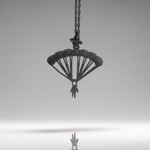May include: A silver pendant necklace with a parachute design. The pendant shows a parachute with a person hanging beneath. The necklace chain is also silver, creating a stylish accessory. The design is detailed.