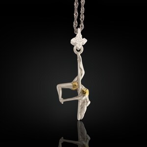 Può includere: Una collana con pendente in argento raffigurante una ballerina in posa da pole dance. Il corpo della ballerina ha accenti dorati. Il pendente è appeso a una catena d'argento. Lo sfondo è nero.