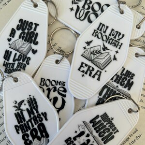 May include: White rectangular keychains with black text and graphics. The keychains have phrases like "Just a Girl in Love with Her Books" and "In My Bookish Era." Each keychain has a silver keyring attached.