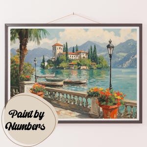 May include: A painting of a picturesque Italian lake scene with a small island featuring a white building and lush greenery. The lake is surrounded by mountains and there are boats on the water. The painting is framed in a dark wood frame and has a "Paint by Numbers" label in the bottom left corner.