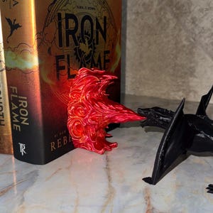 May include: A vibrant red, flame-shaped sculpture and a black dragon figurine are positioned near a book with the title "Iron Flame." The book has a gold cover with the title in bold letters. The scene is set on a marble-like surface.