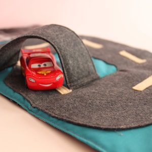 Puede incluir: Un coche de juguete rojo con una cara sonriente conduce a través de un túnel de fieltro gris en una alfombra de juego de fieltro azul y gris.