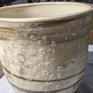 Może przedstawiać: Biała ceramiczna doniczka z brązowymi paskami i kropkowaną glazurą. Doniczka stoi na kole garncarskim.