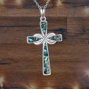 Może przedstawiać: Srebrny naszyjnik z krzyżykiem z inkrustacją z muszli abalonu. Krzyż ma wzór nieskończoności w środku i wisi na delikatnym łańcuszku. Muszla abalonu ma opalizujące odcienie niebieskiego, zielonego i fioletowego.