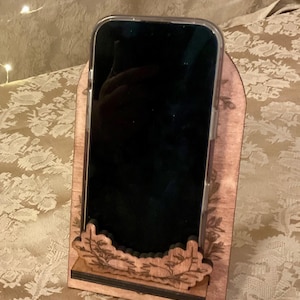 Puede incluir: Un soporte de tel&eacute;fono de madera con un dise&ntilde;o tallado. El soporte es marr&oacute;n y tiene una base negra. Un tel&eacute;fono inteligente est&aacute; en el soporte.