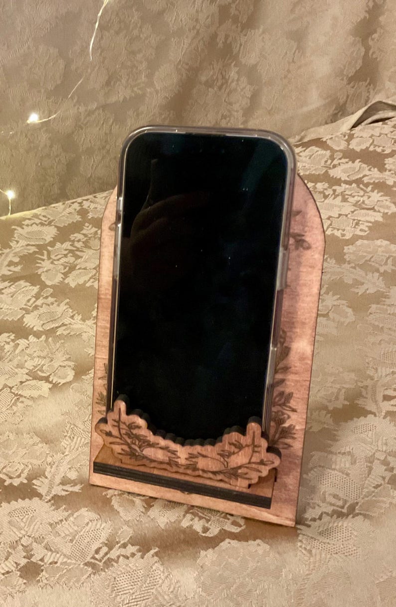 Puede incluir: Un soporte de tel&eacute;fono de madera con un dise&ntilde;o tallado. El soporte es marr&oacute;n y tiene una base negra. Un tel&eacute;fono inteligente est&aacute; en el soporte.