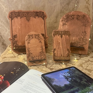 Puede incluir: Tres soportes de tel&eacute;fono de madera de diferentes tama&ntilde;os, cada uno con un dise&ntilde;o floral decorativo. Los soportes est&aacute;n hechos de madera marr&oacute;n claro y tienen un acabado natural. El soporte m&aacute;s grande est&aacute; en el fondo, y el soporte m&aacute;s peque&ntilde;o est&aacute; en primer plano. Una tableta est&aacute; sobre un libro en primer plano.