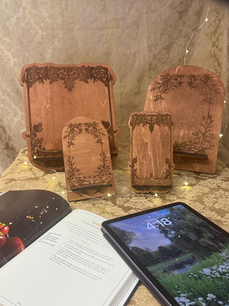 Puede incluir: Tres soportes de tel&eacute;fono de madera de diferentes tama&ntilde;os, cada uno con un dise&ntilde;o floral decorativo. Los soportes est&aacute;n hechos de madera marr&oacute;n claro y tienen un acabado natural. El soporte m&aacute;s grande est&aacute; en el fondo, y el soporte m&aacute;s peque&ntilde;o est&aacute; en primer plano. Una tableta est&aacute; sobre un libro en primer plano.