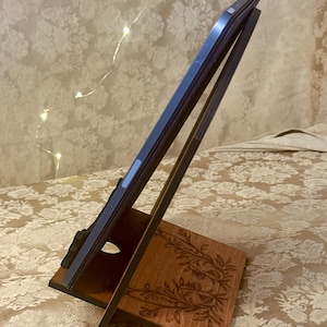 Puede incluir: Un soporte de tableta de madera con un dise&ntilde;o floral. El soporte est&aacute; hecho de madera oscura y tiene un acabado marr&oacute;n claro. El soporte est&aacute; dise&ntilde;ado para sostener una tableta en posici&oacute;n vertical.