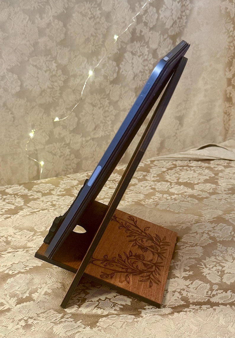 Puede incluir: Un soporte de tableta de madera con un dise&ntilde;o floral. El soporte est&aacute; hecho de madera oscura y tiene un acabado marr&oacute;n claro. El soporte est&aacute; dise&ntilde;ado para sostener una tableta en posici&oacute;n vertical.
