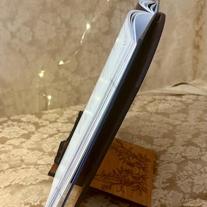 Puede incluir: Un soporte para libros de madera con un dise&ntilde;o floral. El soporte sostiene un libro abierto en una p&aacute;gina con texto. El libro est&aacute; cerrado en el lado izquierdo y abierto en el lado derecho.