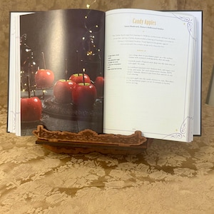 Puede incluir: Un libro de cocina abierto en una p&aacute;gina titulada "Candy Apples" con una receta para hacer manzanas de caramelo. La p&aacute;gina presenta una foto de manzanas de caramelo rojas sobre una superficie negra.
