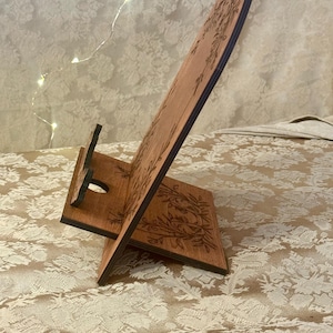 Puede incluir: Soporte de tel&eacute;fono de madera con un dise&ntilde;o floral. El soporte est&aacute; hecho de madera oscura y tiene un recorte para un tel&eacute;fono.