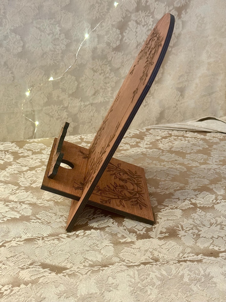 Puede incluir: Soporte de tel&eacute;fono de madera con un dise&ntilde;o floral. El soporte est&aacute; hecho de madera oscura y tiene un recorte para un tel&eacute;fono.