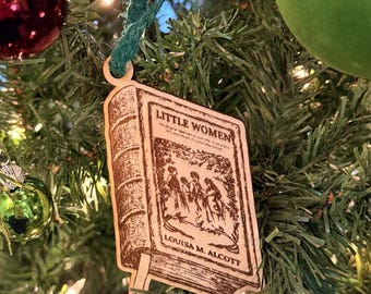 Op de afbeelding: Houten kerstboomversiering in de vorm van een boek, met de titel "Little Women" en de naam van de auteur, Louisa M. Alcott. De versiering hangt aan een groen touwtje en hangt tussen de dennentakken.