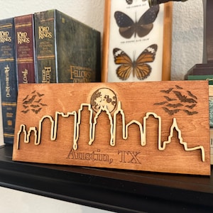 Puede incluir: Un letrero de madera con un diseño de corte por láser del horizonte de Austin, Texas. El letrero presenta una luna llena, murciélagos volando en el cielo y el texto "Austin, TX".