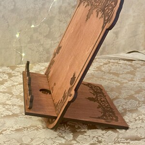 Puede incluir: Un soporte de tel&eacute;fono de madera con un dise&ntilde;o decorativo de un &aacute;rbol y hojas. El soporte est&aacute; hecho de madera marr&oacute;n claro y tiene un recorte para un tel&eacute;fono.