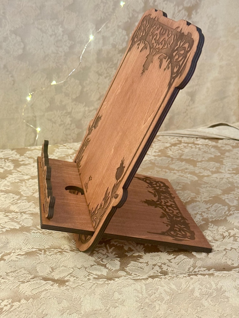 Puede incluir: Un soporte de tel&eacute;fono de madera con un dise&ntilde;o decorativo de un &aacute;rbol y hojas. El soporte est&aacute; hecho de madera marr&oacute;n claro y tiene un recorte para un tel&eacute;fono.