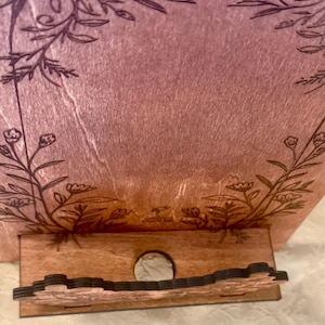 Puede incluir: Un soporte de tel&eacute;fono de madera con un dise&ntilde;o floral. El soporte est&aacute; pintado en un degradado de rosa y morado. El soporte tiene un hueco para un tel&eacute;fono y una peque&ntilde;a repisa para cables de carga.