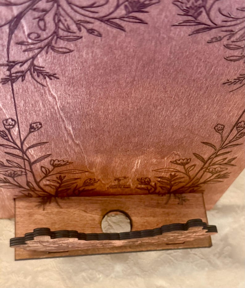 Puede incluir: Un soporte de tel&eacute;fono de madera con un dise&ntilde;o floral. El soporte est&aacute; pintado en un degradado de rosa y morado. El soporte tiene un hueco para un tel&eacute;fono y una peque&ntilde;a repisa para cables de carga.