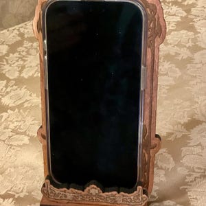 Puede incluir: Un soporte de tel&eacute;fono de madera con tallas intrincadas. El soporte est&aacute; dise&ntilde;ado para sostener un tel&eacute;fono en posici&oacute;n vertical, con una ranura para que el tel&eacute;fono descanse en &eacute;l. El soporte est&aacute; hecho de madera oscura y tiene un acabado natural.