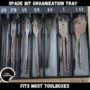 May include: A black plastic tray with slots for organizing spade drill bits. The tray is labeled with different bit sizes: 3/8, 7/16, 1/2, 5/8, 3/4, 1, and 1 1/2. The text "SPADE BIT ORGANIZATION TRAY" is printed at the top of the tray. The text "FITS MOST TOOLBOXES" is printed at the bottom of the tray.