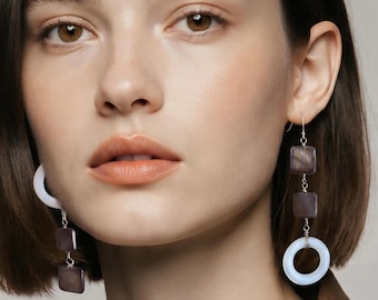 May include: Dangle earrings featuring two dark gray square beads and one smooth white circular ring suspended from a silver-toned hook. The earrings have a vertical design with alternating geometric shapes, showcasing a contrast between the dark square elements and the light circular base.