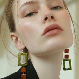 May include: Earrings with a rectangular green frame and a wooden square accent. The earrings are hanging from a silver hook. The earrings are worn by a person wearing a black blazer.