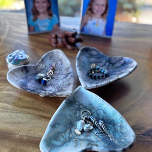 May include: Heart-shaped ceramic jewelry dishes with a marbled blue and gray pattern. Each dish holds various pieces of jewelry, including earrings and rings. Two framed photos of a young girl are in the background.