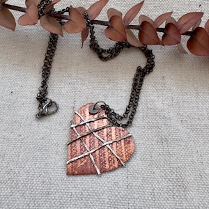 May include: A heart-shaped pendant necklace with a copper-colored base and silver-toned wire accents. The necklace has a dark chain and clasp. The pendant is decorated with a striped pattern. A sprig of dried leaves is in the background.