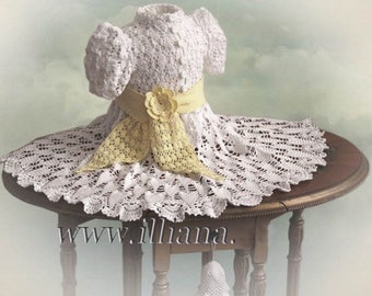 May include: A white crocheted baby dress with a yellow crocheted sash and flower. The dress is displayed on a wooden table with a white crocheted bell hanging from the table.