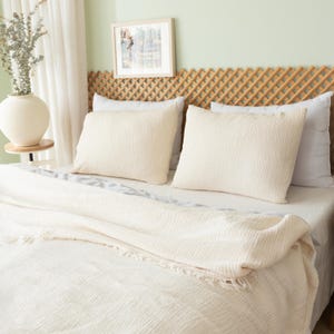 May include: A bed with cream-colored bedding, including a textured blanket and two square pillows. A woven headboard and a framed picture hang above the bed. A white vase with dried stems sits on a small side table.