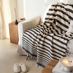 Puede incluir: Una manta a rayas blancas y negras sobre un sofá blanco. La manta tiene un borde con flecos y está hecha de tela tejida. La habitación tiene una paleta de colores neutros con una mesa auxiliar de madera y un par de zapatillas en el suelo.