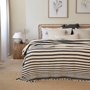 Op de afbeelding: Een bed is bedekt met een zwart-crème gestreepte deken. De deken heeft een franje langs de onderrand. Het bed heeft een geweven hoofdbord en is versierd met witte kussens. Een houten nachtkastje met een lamp staat naast het bed.