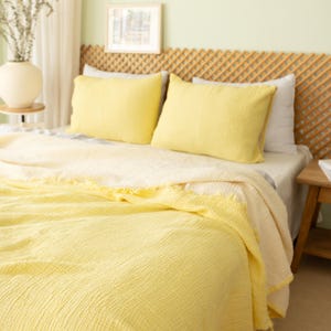 Puede incluir: Una cama con una manta amarilla y dos almohadas amarillas. La cama tiene un cabecero tejido y una pequeña mesita de noche de madera. Un jarrón con flores secas está sobre una mesita. La habitación tiene una pared verde claro.