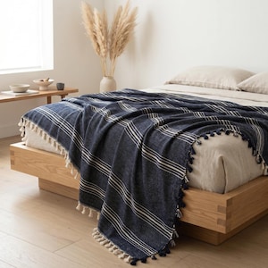 May include: A navy blue throw blanket with white horizontal stripes and decorative tassels along the edges, draped over a light-coloured bed. The blanket features a textured weave pattern and is positioned on a wooden bed frame in a bright room.