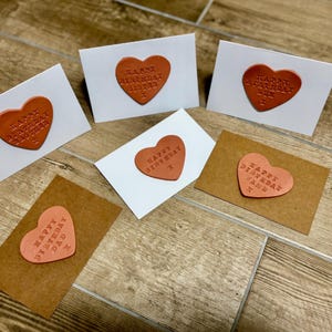 May include: A collection of birthday cards with heart-shaped embellishments. The cards feature embossed text that reads "Happy Birthday" along with names like "Brother" and "Dad." The hearts are in shades of orange and pink, set against white and brown cardstock.