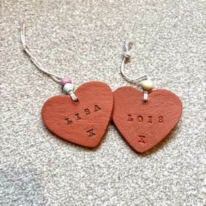 May include: Two terracotta heart-shaped decorations, each bearing a name: "LISA" and "LOIS". The names are stamped, with an "X" below each. They hang from white cords with wooden beads, ideal for gifting or home decor.